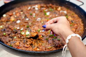 A hand stirs a colorful seafood paella filled with fresh ingredients under the warm summer sun at a lively beach gathering, filled with laughter and joy.